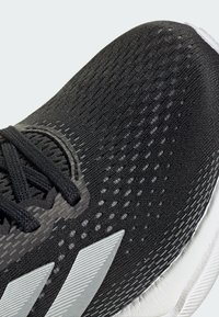 Chaussure de sport noire avec une tige en tissu texturé comportant des zones en mesh, des rayures d'accent gris clair et une semelle blanche.