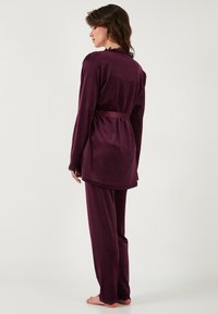 Woman standing barefoot wearing dark purple velvet long-sleeve top with belt and matching pants, shown from back against plain background.