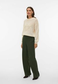 Kremowy, dziergany sweter o luźnym kroju z teksturowanym emblematem. Połączony z zielonymi, szerokimi spodniami z wysokim stanem oraz ciemnymi szpilkami.