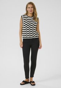 Haut sans manches en tricot chevron noir et blanc associé avec un jean skinny noir et des sandales noires sur un fond uni.