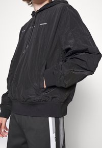 Veste en nylon noir avec capuche, dotée de poignets élastiques, d'une poche avant et d'un texte logo blanc sur la poitrine. Matériau léger et texturé.