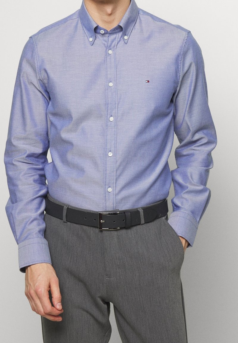 Camicia azzurro chiaro con colletto, caratterizzata da una texture sottile e bottoni bianchi, abbinata a pantaloni grigi e una cintura nera.