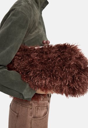 Osoba ubrana w ciemnozieloną kurtkę i brązowe spodnie trzyma dużą, puszystą, o kręconej fakturze brązową torebkę z okrągłymi klamrami.