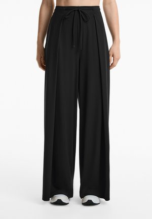 Pantaloni neri a gamba larga con vita regolabile, realizzati in tessuto morbido, caratterizzati da pences e indossati con sneakers bianche e nere.