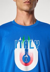T-shirt en coton bleu avec un design graphique portant le mot "ITALIE", souligné de blanc et d'accents verts, et un logo Adidas en bas.