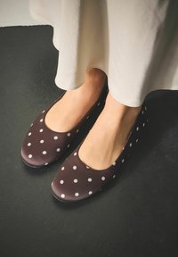 Brązowe baleriny w groszki o gładkiej fakturze i zaokrąglonym nosku, zestawione z jasną, zwiewną spódnicą. Czarny tle. 