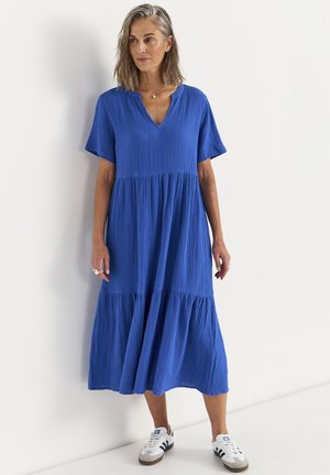 Kvinde med skulderlangt gråt hår står iført en løs blå midi-kjole med V-hals og hvide sneakers med sorte striber.