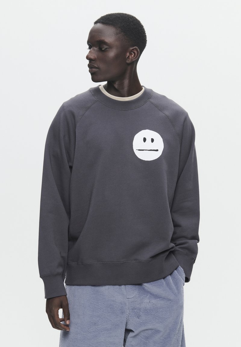 Grau Baumwollpullover mit einem runden, weißen Gesichtsgrafik auf der Brust. Mit Rundhalsausschnitt und gerippten Bündchen. Lockere Passform.
