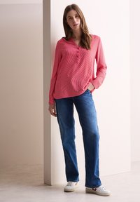 Camicetta a maniche lunghe a righe rosa chiaro con colletto abbottonato. Indossata con jeans blu in denim e sneaker bianche, in piedi contro uno sfondo neutro.