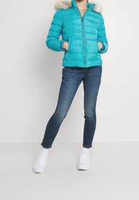 Turquoise pufferjack met een met bont afgewerkte capuchon, ritssluiting en twee zijzakken, gecombineerd met donkerblauwe strakke jeans en witte sneakers.