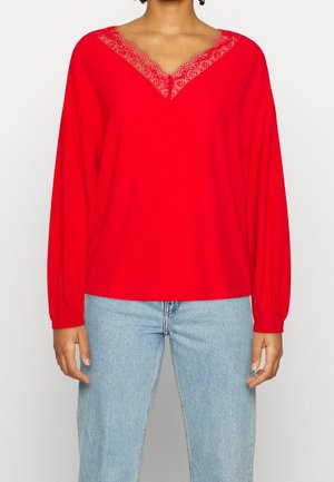 Kobieta ubrana w czerwoną bluzkę z długim rękawem z koronkowym dekoltem w szpic, w połączeniu z jasnoniebieskimi jeansami, stojąca na jednolitym tle.