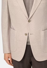 Blazer beige clair avec une texture douce, fermeture à un bouton, revers crantés, et deux poches avant, associé à un pull gris et un pantalon noir.
