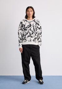 Knit sweater in white with black abstract patterns, round neckline, ribbed cuffs, paired with black pants and sneakers. Plain background.