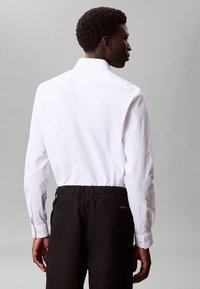 Hvid langærmet skjorte med klassisk krave, kombineret med sorte bukser med elastik i taljen. Glat stof og skræddersyet pasform synlig bagfra.