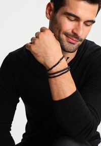 Svarte perle- og flettede armbånd som bæres på håndleddet, med forskjellige teksturer og former, mot en bakgrunn av en svart skjorte.