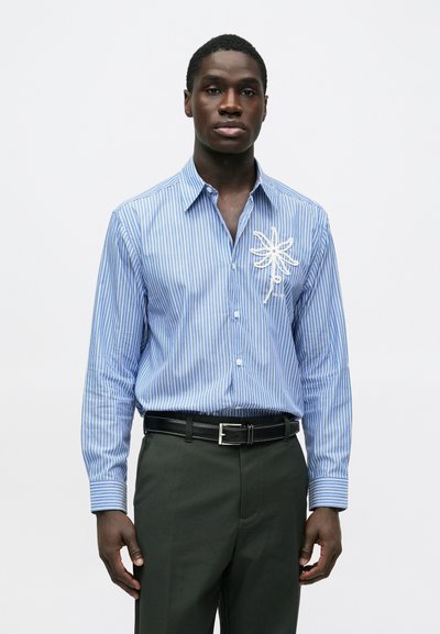 Homme portant une chemise boutonnée à rayures bleues et blanches avec une fleur brodée blanche sur la poitrine, ceinture noire et pantalon sombre.