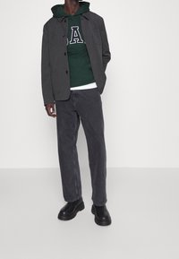 Grijze jas met knopen, groene hoodie met een letterlogo, gelaagd over een witte shirt, gecombineerd met donkergrijze jeans en zwarte laarzen.