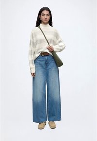 Kvinde iført en hvid stribet sweater, brede blå jeans, brune loafers og en grøn crossbody-taske mod en enkel hvid baggrund.