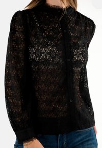 Blusa nera in pizzo con motivi a diamante intricati, chiusura a bottoni e colletto alto con bordo ondulato. Maniche lunghe, leggermente gonfie.