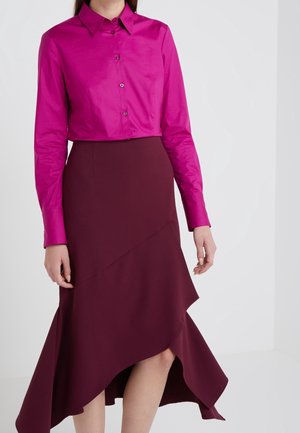 Femme portant une chemise boutonnée magenta vif et une jupe asymétrique bordeaux foncé sur un fond clair uni.