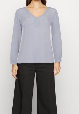 Femme portant un chemisier bleu clair à manches longues et col en V, associé à un pantalon noir ample, debout devant un fond uni.