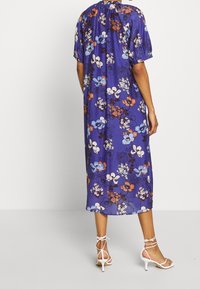 Bloemige midi-jurk in blauw met korte mouwen, voorzien van levendige, meerkleurige bloemenpatronen en een losse snit. Gecombineerd met witte sandalen met bandjes.