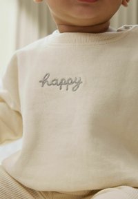 Lys beige bomulds-sweatshirt med grå broderet tekst, der står "happy." Blød tekstur med ribbet rund hals og manchetter.