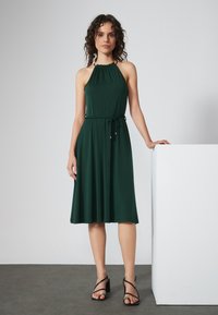Green halter neckline dress with an elastic waist and a drawstring. Smooth fabric, flowing skirt, paired with black strappy sandals.