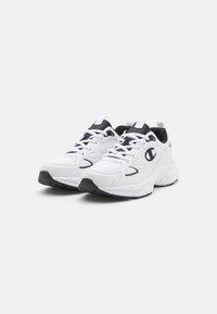 Pari valkoisia ja mustia urheilutennareita, joissa on verkkopaneelit, nauhat ja paksut pohjat, asetettu vierekkäin valkoista taustaa vasten.