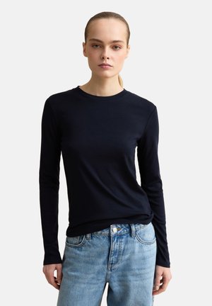 Donna con una maglietta nera aderente a maniche lunghe e jeans azzurri a vita alta, in piedi davanti a uno sfondo chiaro e semplice.