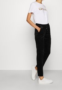 T-shirt blanc à manches courtes avec un motif graphique doré et noir, associé à un jogging en velours noir avec poches latérales et cordon de serrage.