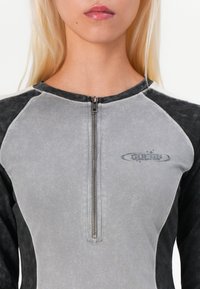 Sivi i crni zip-up dukserica s visokim ovratnikom, teksturiranim materijalom, raglan rukavima i logom na grudima. Sadrži metalni patent zatvarač.