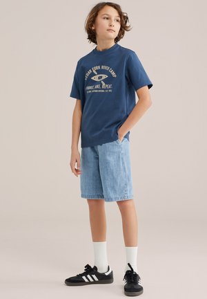 Pois seisab käed taskutes, seljas sinise graafilise kujundusega T-särk, helerohelised teksapüksid, valged sokid ja mustad tossud, ühtlase taustaga.