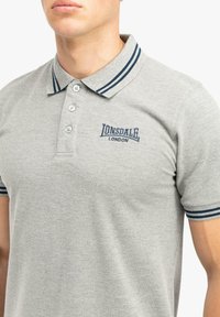 Grå bomulds polo shirt med en tre-knaps placket, krave med blå striber og broderet "Lonsdale London" logo på brystet.