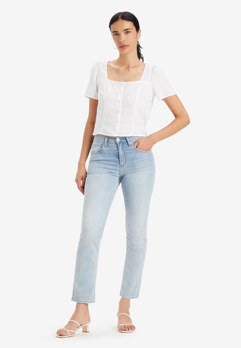 Blouse blanche courte avec décolleté carré et manches bouffantes courtes, associée à un jean taille haute bleu clair et des talons blancs à lanières.