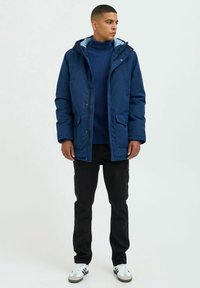 Navy insulated jacket with a zip and button closure, two front pockets, and a hood, layered over a dark blue ribbed sweater and black pants.