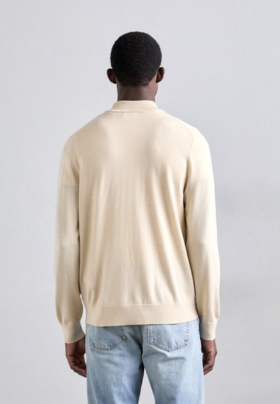 Beige gestrickter Pullover mit klassischem Kragen und langen Ärmeln, der eine glatte Textur und ein schlichtes Design aufweist. Über hellblauen Jeans getragen.