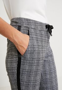 Szare spodnie w kratę z gładkiego materiału, z czarnymi akcentami po bokach i kokardką w talii, oraz bocznymi kieszeniami.