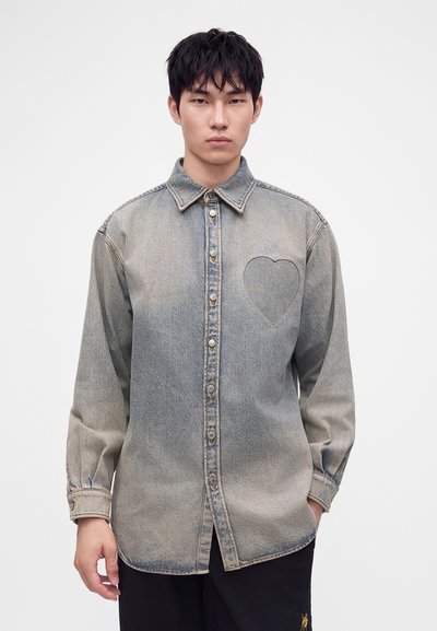 Jeune homme portant une chemise en jean grise délavée à boutons avec un contour subtil en forme de cœur sur la poitrine, debout devant un fond uni.