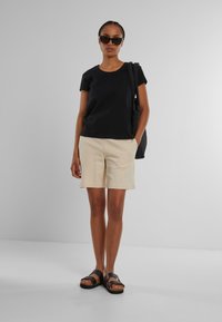 Svart kortermet t-skjorte, beige shorts og svarte sandaler med treglass. Modellen bærer en svart veske. Nøytral bakgrunn.