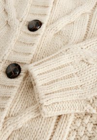 Cream-colored knit cardigan featuring thick cables and ribbed textures, with large dark buttons and a folded cuff detail.