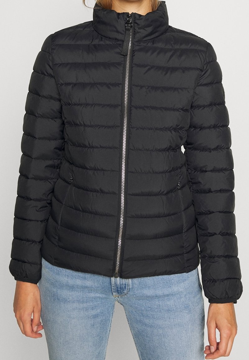 Veste matelassée noire avec fermeture éclair à l'avant, col montant, poches latérales portée par une personne en jean bleu clair, raccourcie au niveau du cou et des hanches.