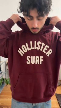 Młody mężczyzna poprawiający bordową bluzę z kapturem z napisem "HOLLISTER SURF" w jasnym pokoju z drewnianą podłogą i roślinami doniczkowymi.