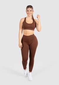 Bruine sportieve crop top en high-waisted leggings, gemaakt van rekbare stof. Heeft een naadloos ontwerp en een subtiel logo. Witte sneakers maken de look compleet.