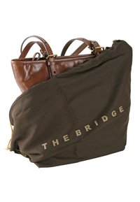 Borsa in pelle marrone parzialmente coperta da un sacchetto protettivo marrone scuro con la scritta "THE BRIDGE" in lettere maiuscole.