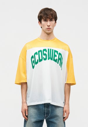 Νεαρός άνδρας με φαρδύ κίτρινο και άσπρο πουκάμισο από δίχτυ με πράσινο κείμενο "GCDSWEAR", συνδυασμένο με μπλε τζιν, στέκεται μπροστά σε απλό φόντο.