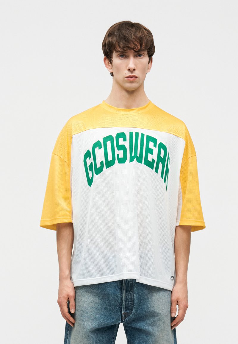 Νεαρός άνδρας με φαρδύ κίτρινο και άσπρο πουκάμισο από δίχτυ με πράσινο κείμενο "GCDSWEAR", συνδυασμένο με μπλε τζιν, στέκεται μπροστά σε απλό φόντο.