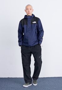 Chaqueta de lluvia marina con acentos negros, que cuenta con cierre de cremallera y capucha, combinada con pantalones negros que tienen detalles reflectantes y un corte relajado.