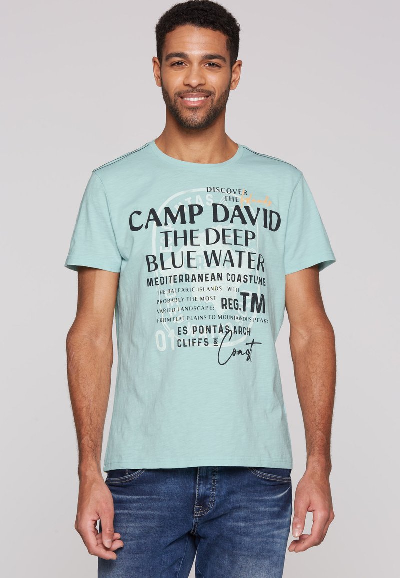 Světlá tyrkysová tričko s kulatým výstřihem, krátkými rukávy a výrazným černým grafickým textem s vícebarevným akcentem na přední straně.