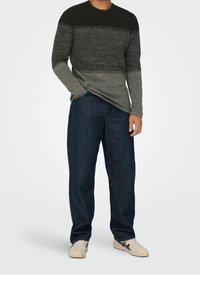 Langärmliges Strickoberteil mit einem Farbverlaufsmuster in Schwarz, Grau und Dunkelgrau, kombiniert mit locker sitzenden dunkelblauen Denim-Jeans und weißen Sneakern.